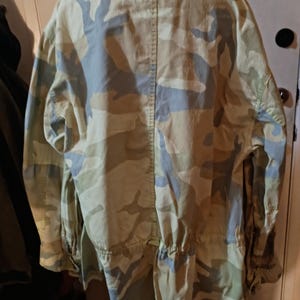 K&ouml;nnte beinhalten: Eine Tarnjacke in Beige-, Gr&uuml;n- und Blaut&ouml;nen. Die Jacke hat einen Kragen, lange &Auml;rmel und eine Taille mit Kordelzug. Die R&uuml;ckseite der Jacke ist sichtbar und zeigt das Tarnmuster.