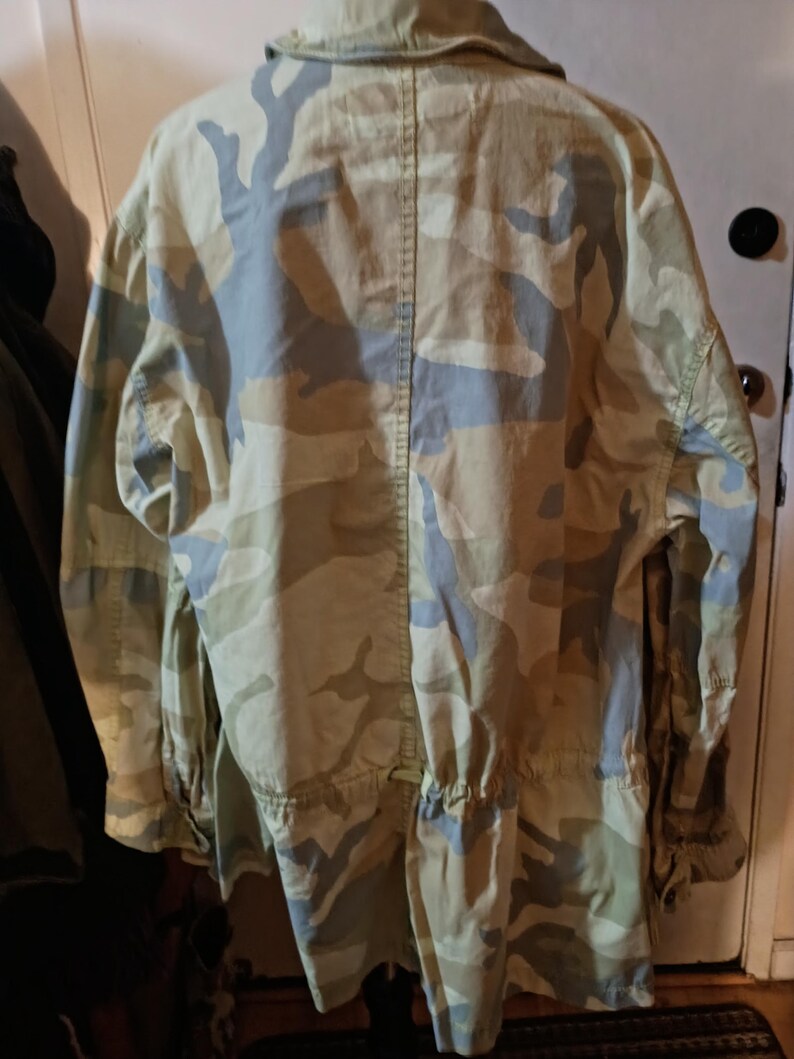 K&ouml;nnte beinhalten: Eine Tarnjacke in Beige-, Gr&uuml;n- und Blaut&ouml;nen. Die Jacke hat einen Kragen, lange &Auml;rmel und eine Taille mit Kordelzug. Die R&uuml;ckseite der Jacke ist sichtbar und zeigt das Tarnmuster.