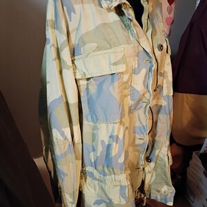 K&ouml;nnte beinhalten: Eine Tarnjacke in Hellgr&uuml;n, Blau und Beige. Die Jacke hat einen Kragen, einen Rei&szlig;verschluss und mehrere Taschen. Der Stoff hat eine leicht zerknitterte Textur und die Jacke wirkt leicht.