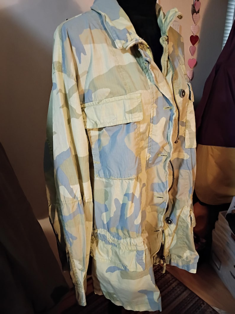 K&ouml;nnte beinhalten: Eine Tarnjacke in Hellgr&uuml;n, Blau und Beige. Die Jacke hat einen Kragen, einen Rei&szlig;verschluss und mehrere Taschen. Der Stoff hat eine leicht zerknitterte Textur und die Jacke wirkt leicht.