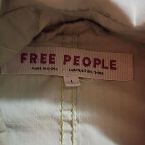 K&ouml;nnte beinhalten: Nahaufnahme eines hellgr&uuml;nen Kleidungsst&uuml;cks mit einem gewebten Etikett mit der Aufschrift "FREE PEOPLE" in roten Buchstaben. Das Etikett enth&auml;lt auch die Angaben "MADE IN CHINA" und "FABRIQUE EN CHINE". Das Kleidungsst&uuml;ck hat ein Gr&ouml;&szlig;enetikett "L".