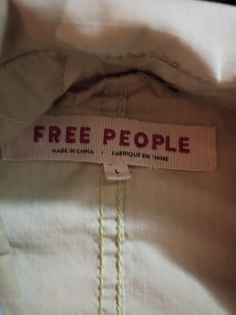 K&ouml;nnte beinhalten: Nahaufnahme eines hellgr&uuml;nen Kleidungsst&uuml;cks mit einem gewebten Etikett mit der Aufschrift "FREE PEOPLE" in roten Buchstaben. Das Etikett enth&auml;lt auch die Angaben "MADE IN CHINA" und "FABRIQUE EN CHINE". Das Kleidungsst&uuml;ck hat ein Gr&ouml;&szlig;enetikett "L".
