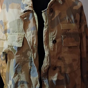 K&ouml;nnte beinhalten: Tarnmuster-Jacke in Beige-, Olivgr&uuml;n- und Graut&ouml;nen. Die Jacke hat einen Rei&szlig;verschluss, mehrere Taschen und Kn&ouml;pfe. Der Kragen ist im klassischen Stil.