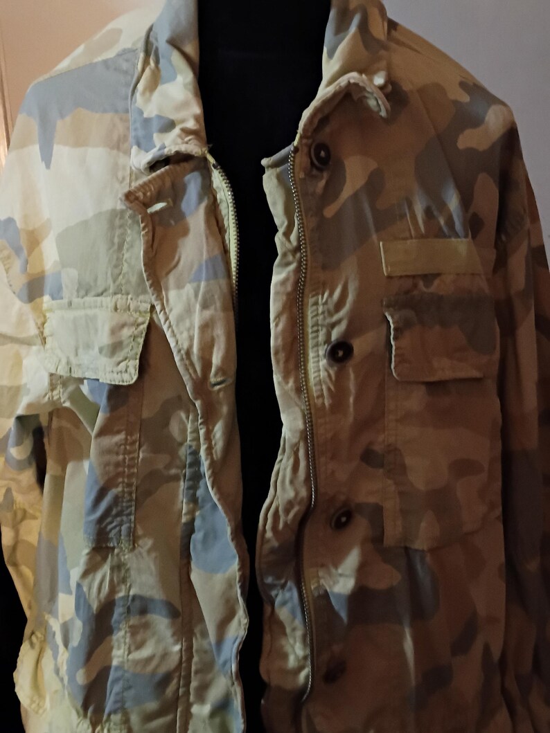 K&ouml;nnte beinhalten: Tarnmuster-Jacke in Beige-, Olivgr&uuml;n- und Graut&ouml;nen. Die Jacke hat einen Rei&szlig;verschluss, mehrere Taschen und Kn&ouml;pfe. Der Kragen ist im klassischen Stil.
