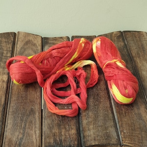 Puede incluir: Dos ovillos de hilo rojo y amarillo teñido anudado sobre una superficie de madera.