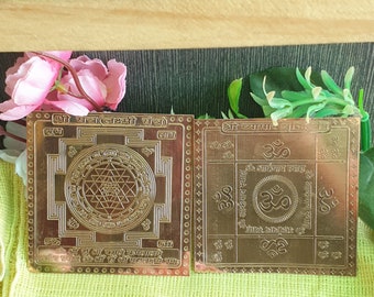 Puede incluir: Dos yantras de metal de color dorado, uno con un diseño de Lakshmi Yantra y el otro con un diseño de Shri Yantra. Ambos yantras están rodeados por un borde decorativo y presentan texto sánscrito.