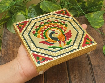 Può includere: Una scatola decorativa quadrata color oro con un disegno di pavone. Il pavone è raffigurato con piume verdi, rosse e gialle vibranti. La scatola ha un bordo geometrico e piccoli piedini bianchi.