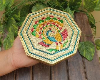 May include: Octagonal gold-coloured box with a peacock design. The peacock is painted in shades of green, blue, yellow, and red. The box has a decorative border.