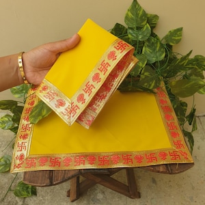 Könnte beinhalten: Zwei gelbe Tischsets mit einem rot-goldenen Rand mit sich wiederholendem Design. Ein Tischset ist auf dem anderen gestapelt. Die Tischsets stehen auf einem kleinen Holztisch, mit grünen Blättern im Hintergrund.