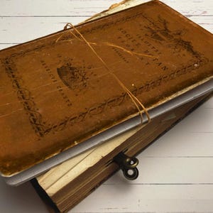 May include: An antique book with a brown leather cover, secured with twine. The cover displays decorative text and an emblem. The book's pages are aged, and the edges show wear. The book is stacked on top of another book with a clasp.
