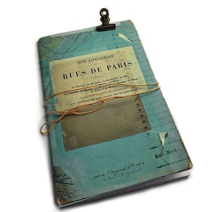 May include: A blue and white vintage notebook with a brown leather strap and a brass binder clip. The cover has the text "Liste Alphabetique des Rues de Paris" and "Paris, August 9th 1890".