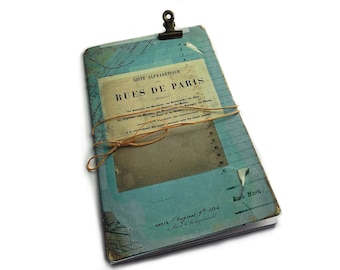 May include: A blue and white vintage notebook with a brown leather strap and a brass binder clip. The cover has the text "Liste Alphabetique des Rues de Paris" and "Paris, August 9th 1890".