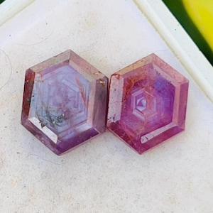 Puede incluir: Dos gemas hexagonales facetadas con un tono púrpura rojizo. Las gemas están sobre una superficie blanca, con un fondo borroso verde y amarillo. Las gemas tienen patrones internos y probablemente son para la fabricación de joyas.