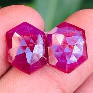 Peut inclure: Deux rubis taillés en hexagone avec une couleur rouge magenta profond et des surfaces facettées. Les pierres précieuses présentent un haut degré de clarté et une réflexion de la lumière sur leurs formes géométriques à six côtés.