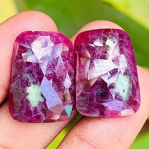 May include: Two faceted, deep red gemstones with a mix of purple and green hues. The gems have a rectangular shape with a rounded top. The gems are held in a hand against a blurred green background.