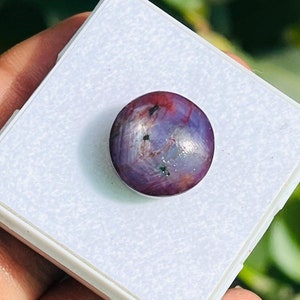 May include: A round, polished gemstone with a mix of purple and red hues, set on a white square. The gemstone has a smooth surface with subtle variations in color and texture, suggesting it is a natural stone.