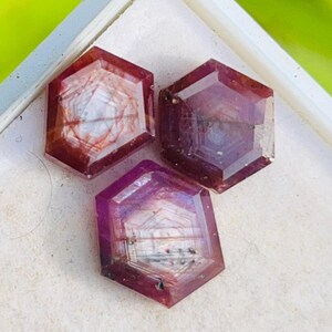 Może przedstawiać: Trzy sześciokątne kamienie szlachetne o głębokim czerwono-fioletowym odcieniu są prezentowane w białej tacce. Kamienie mają wewnętrzne wzory i fasetki, które odbijają światło. Tło jest miękkie, rozmyte zielone.