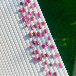 May include: A collection of small, raw, unpolished pink and reddish gemstones arranged on a white, ridged, rectangular tray. The stones vary in shape and size, displaying natural textures and crystalline surfaces with deep magenta and ruby hues.