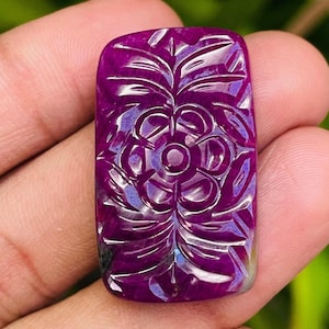 Może przedstawiać: Prostokątny, rzeźbiony kamień szlachetny w odcieniach głębokiej magenty. Kamień szlachetny ma kwiatowy wzór ze skomplikowanymi detalami. Rzeźba jest wyśrodkowana na kamieniu, z kwiatem pośrodku i otaczającymi go liśćmi. Kamień szlachetny jest trzymany w dłoni.