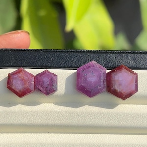 以下が含まれることがあります： 黒いディスプレイケースに入った4つの六角形の宝石。宝石は赤と紫のさまざまな色合いです。宝石はファセット加工されており、研磨されているように見えます。ディスプレイケースは白い内装です。