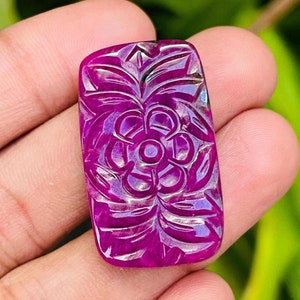 May include: A rectangular, carved gemstone in a deep magenta hue. The gemstone features a floral design with detailed petals and leaves. The carving is intricate, with light reflecting off the polished surfaces. The gemstone is held in a hand, with a blurred green background.