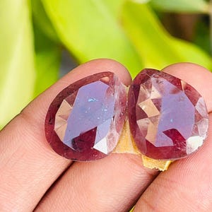 May include: Two pear-shaped, faceted gemstones with a deep, translucent red colour. The stones feature a polished surface with multiple geometric facets that reflect light, showing internal dark inclusions and a smooth, rounded teardrop silhouette.