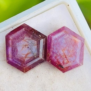 以下が含まれることがあります： ピンクと紫の色合いの、六角形のファセット宝石が2つ。白いトレイにセットされ、背景はぼやけた緑色。石には内部のインクルージョンがあり、研磨された表面が特徴です。