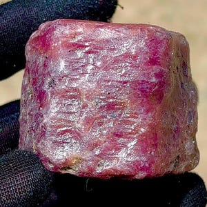 May include: A raw, unpolished ruby crystal with a deep magenta-red hue. The gemstone features a rough, textured surface with natural crystalline facets and opaque sections, displaying a cylindrical shape with irregular edges and a dense, solid structure.