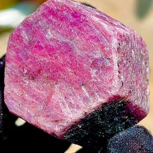 May include: A raw, hexagonal ruby crystal displaying a vibrant magenta-pink hue with a textured, crystalline surface. The gemstone features natural striations and facets across its top face, showcasing a deep, saturated colour throughout the stone.