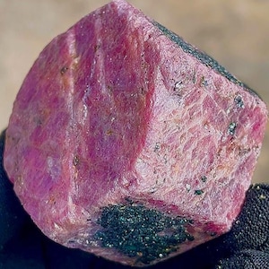 Pode incluir: Um rubi bruto, sem corte, com um tom rosa profundo. A superfície tem uma aparência texturizada com pequenas inclusões verde-escuras. O rubi é segurado em uma luva preta. Uma gema natural para joalheria.