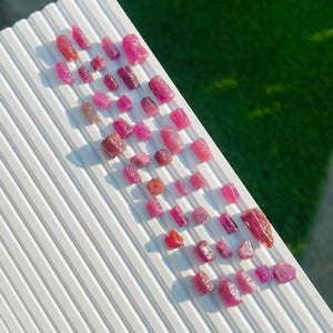 Pode incluir: Uma coleção de pedras preciosas de rubi bruto em várias formas e tamanhos, dispostas sobre uma superfície branca e estriada. Os rubis exibem tons de rosa e vermelho, alguns parecendo translúcidos. O fundo inclui uma mancha verde.