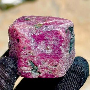 May include: A raw, unpolished ruby gemstone with a deep magenta and pink hue. The stone features a blocky, crystalline structure with visible natural inclusions and dark green mineral deposits embedded within its textured, multifaceted surface.
