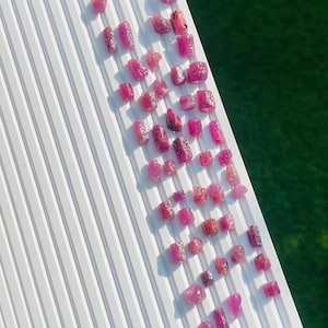 May include: A collection of small, raw, pinkish-red gemstone crystals arranged in rows on a white, ridged display tray. The stones vary in shape and size, exhibiting natural textures and translucent to opaque qualities across the group.