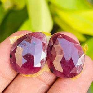 May include: Two faceted, pear-shaped rubies with a deep magenta-red hue. Each gemstone features a polished surface with multiple geometric facets that reflect light, showcasing a translucent quality and natural inclusions within the stones.