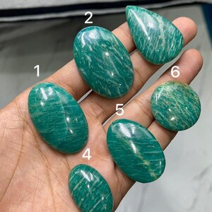 Puede incluir: Seis cabujones de amazonita pulida de diversas formas y tamaños, etiquetados con los números del uno al seis. Cada piedra presenta un vibrante color verde azulado con inclusiones blancas nubosas características y una superficie lisa, brillante y reflectante.