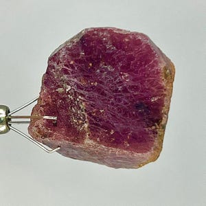 May include: A raw, deep magenta-coloured gemstone with a rough, crystalline texture and irregular edges. The stone is held by a small, silver-toned metal wire clip that grips the side, showcasing its natural, unpolished surface and dark red hues.