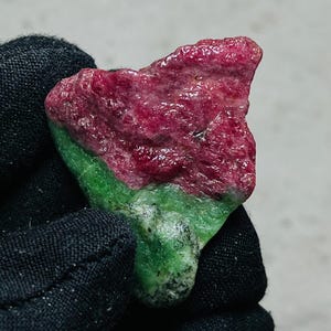 Könnte beinhalten: Ein rohes, unpoliertes Mineralexemplar mit einer deutlichen Farbtrennung. Der obere Teil ist tief, leuchtend magentarot mit einer kristallinen, strukturierten Oberfläche, während der untere Teil ein massives, undurchsichtiges Waldgrün ist.