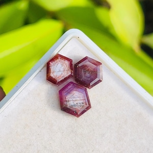 Puede incluir: Tres gemas hexagonales de color púrpura rojizo se exhiben en una bandeja blanca. Las gemas tienen patrones internos y se colocan sobre un fondo verde borroso. Las gemas son probablemente para la fabricación de joyas.