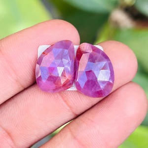 May include: Two faceted, teardrop-shaped gemstones in shades of pink and purple. The gems are held on a white backing, showcasing their cut and color. The stones are held in a hand, with a blurred green background.