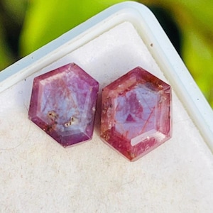 以下が含まれることがあります： 深いピンク色の六角形のファセット宝石が2つ。内部に模様があります。宝石は白い背景に置かれ、その透明度と色を際立たせています。石は研磨仕上げで、光を反射します。