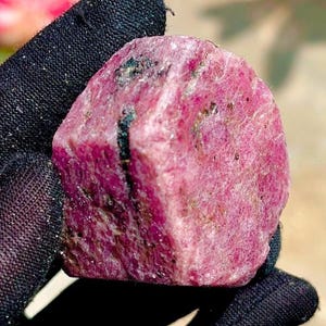 May include: A raw, uncut ruby gemstone with a deep magenta and pink hue. The stone features a rough, crystalline texture with visible natural inclusions and dark mineral deposits running through the surface, held against a black fabric background.