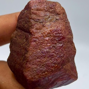 May include: A raw, unpolished ruby gemstone with a deep, dark red color and a textured, uneven surface. The stone features natural crystalline formations and a matte finish, displaying variations in tone across its irregular, blocky shape.