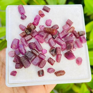 Może przedstawiać: Kolekcja surowych kamieni szlachetnych rubinu o różnych kształtach i rozmiarach, od jasnoróżowych po ciemnobrązowo-czerwone odcienie. Kamienie szlachetne są prezentowane na białej powierzchni, z zielonymi liśćmi w tle.