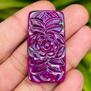 Puede incluir: Una gema rectangular de color magenta intenso con un diseño floral tallado. La intrincada talla presenta una flor central rodeada de hojas y pétalos estilizados. La superficie pulida refleja la luz, resaltando los detalles del diseño.