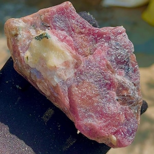 Puede incluir: Una gema en bruto, sin cortar, de color rosa vibrante, con una mancha blanca cremosa y motas verdes. La superficie de la piedra tiene un aspecto texturizado, mostrando formaciones minerales naturales. La gema se sostiene en un guante negro.