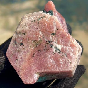May include: A raw, multifaceted mineral specimen featuring a prominent dusty rose and pale pink coloration. The stone displays a crystalline, textured surface with visible internal fractures and small, dark green to black mineral inclusions throughout.