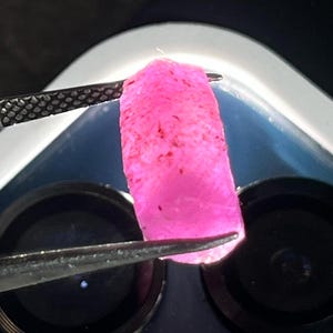 May include: A vibrant pink gemstone, held by tweezers, is the focal point. The gem has a rough, textured surface with dark red inclusions. The lighting highlights the stone's color and internal structure. The background is a blurred view of a microscope.