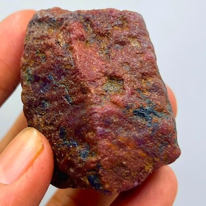 May include: A raw, unpolished gemstone with a rough, textured surface. The stone displays a deep reddish-maroon base colour with visible inclusions of dark blue and metallic gold-toned flecks scattered across its irregular, blocky shape.