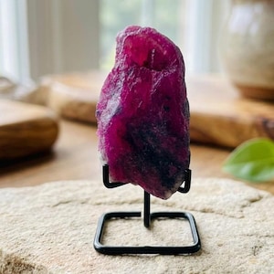 Peut inclure: Une pierre précieuse de rubis brute, principalement magenta avec des inclusions violet foncé et noires, montée sur un petit support en métal noir. La pierre brute est exposée sur une surface neutre, mettant en valeur sa texture naturelle et ses variations de couleur.