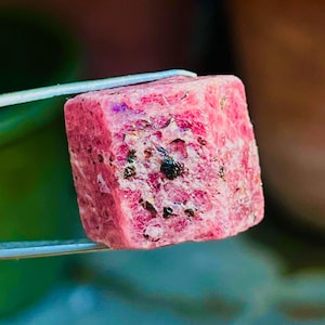 Puede incluir: Primer plano de una gema rosa en bruto sujeta con pinzas. La gema tiene forma de cubo con una superficie texturizada y motas negras y de otros colores. El fondo está borroso, con tonos verdes y marrones.